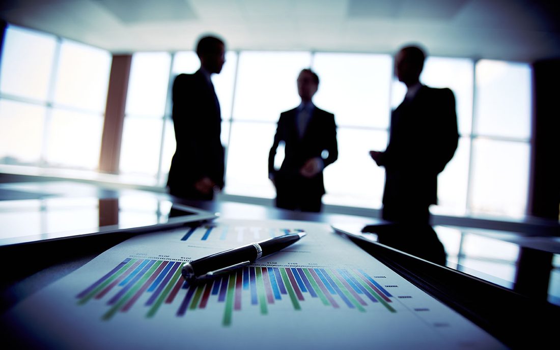 Gráficas financieras impresas y una pluma en primer plano, con tres ejecutivos conversando a contraluz al fondo